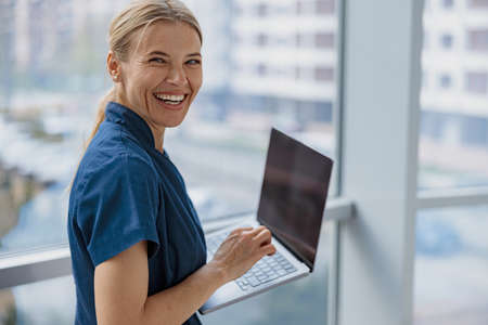 Smiling female healthcare worker using laptop while working at doctors office and looking camera