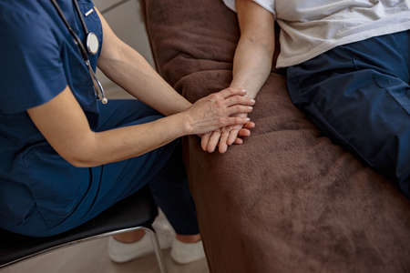 Doctor holding a sick patient by hand supporting her sitting in a hospital roomの写真素材
