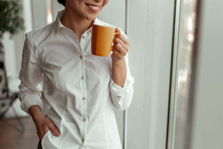 Close up of smiling asian female worker drinking coffee standing near windows at officeの写真素材