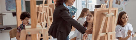 Young beautiful confident teacher is helping a kid to draw on a group lesson in a white modern...