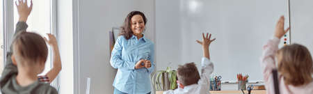 Young happy elementary school pupils at light modern classroom are rising their hands on a lesson