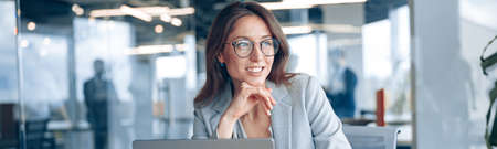 Smiling businesswoman with glasses working on laptop and looking out the window