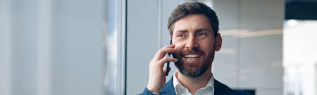 Smiling businessman uses mobile phone to call his business partner while working in the officeの写真素材