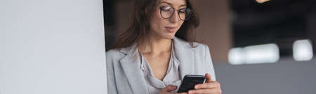 Focused Businesswoman wearing glasses with mobile phone in hand standing in modern officeの写真素材