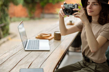 Smiling female traveler using photo camera while sitting in cafe outdoorsの写真素材