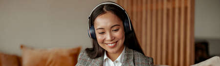 Attractive asian business woman listening music on headphones sitting at laptop with coffee