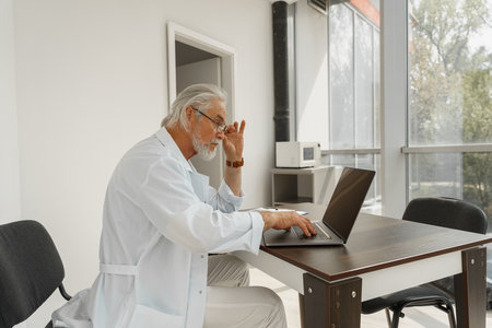 Senior healthcare worker in glasses using laptop while working at doctors officeの写真素材