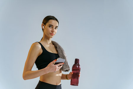 Healthy sporty woman standing with water bottle and phone over studio background