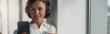 Smiling Business woman holding cup of coffee and using phone while standing near window at officeの写真素材