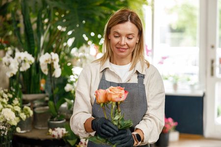 Smiling woman florist hold roses ready to sell on floral background and looking at flowers
