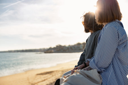 Beautiful romantic couple sitting at the beach wearing casual clothes and looking at the oceanの写真素材