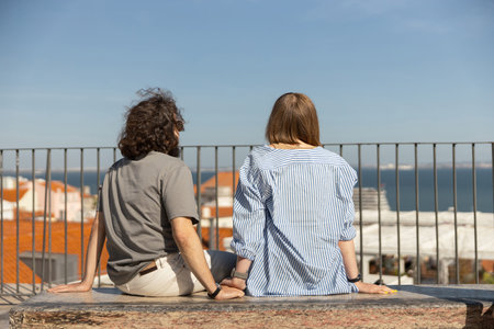 Rear view of young couple is hugging while sitting on bench and enjoys beautiful ocean viewの写真素材