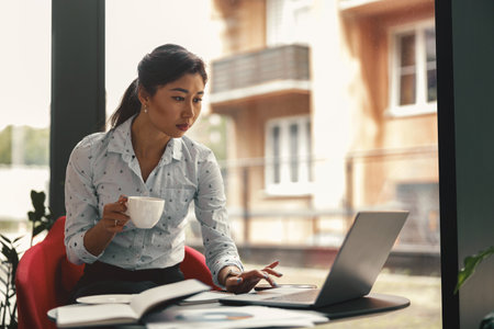 Female entrepreneur working on laptop and drinking coffee while sitting the desk in modern coworkingの写真素材