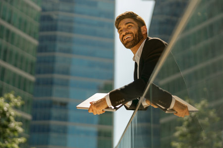 Stylish smiling businessman in suit is standing with laptop on office terrace and looks awayの写真素材