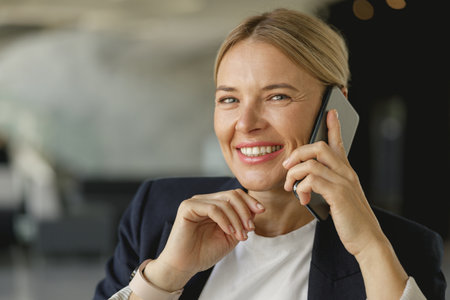 Smiling female entrepreneur talking phone sitting on modern office background and looks cameraの写真素材