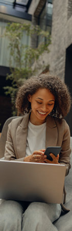 Smiling female manager holding mobile phone while sitting in office and work on laptopの写真素材