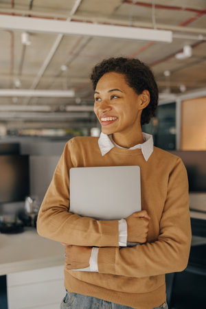 Smiling woman office worker with laptop standing on background of modern office and looks away