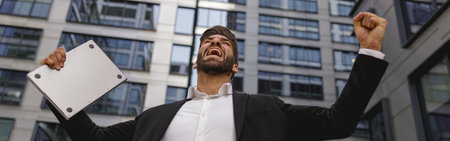 Businessman with laptop standing on office terrace and raised fist is suttisfied of finished projectの写真素材
