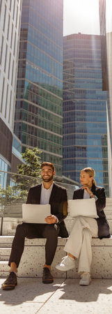 Man and woman in classic suit discussing business details and using laptop while sitting outdoors