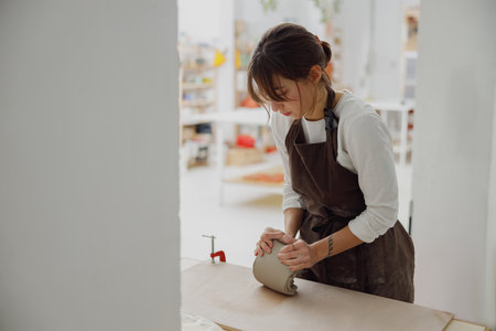 Professional female potter in apron kneads piece of clay with her hands on table in studioの写真素材