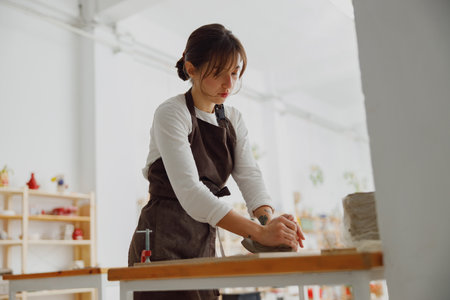 Professional female potter in apron kneads piece of clay with her hands on table in studio