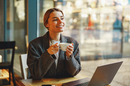 Young female freelancer is drinking coffee in cafe while working on laptop near windowの写真素材