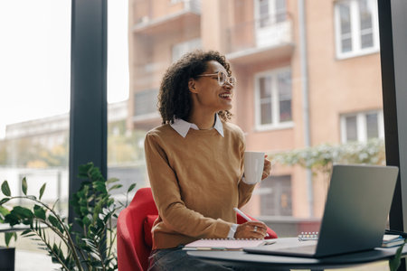 Smiling female student working on laptop and making notes while drinking coffee in cozy coworkingの写真素材