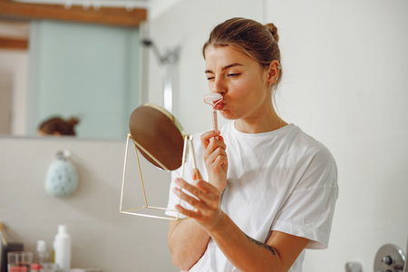 Smiling woman massaging her face with jade roller massager and looking at round cosmetic mirror