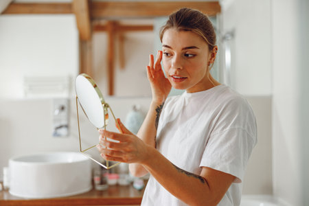 Young woman looking in small round mirror and checking skin while standing in bathroomの写真素材