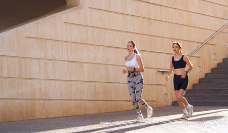 Two young women in sportswear are running on modern buildings background. Active lifestyle conceptの写真素材