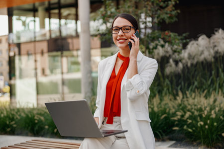 A dedicated businesswoman works outdoors on her laptop while on a phone call, showing commitment