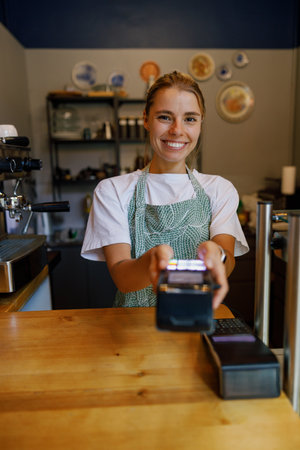 A smiling barista at a cozy coffee shop, ready with a card reader to serve customersの写真素材