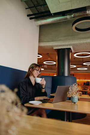 A woman works diligently in a cozy modern cafe with her laptop and warm coffee nearbyの写真素材