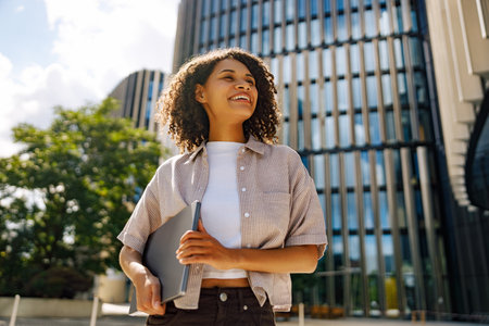 A Confident Young Woman is Joyfully Smiling with Her Laptop in a Modern Urban Settingの写真素材