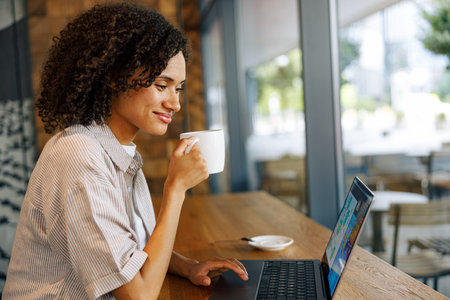 A Woman is Enjoying Her Coffee While Working on Her Laptop in a Modern Cafe Settingの写真素材