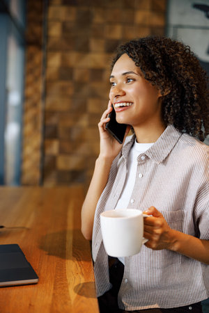 A Woman Relishing Her Coffee While Engaging in a Phone Conversation in a Cozy Cafeの写真素材
