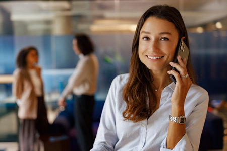 A professional woman is seen smiling while using her phone in a modern office environmentの写真素材
