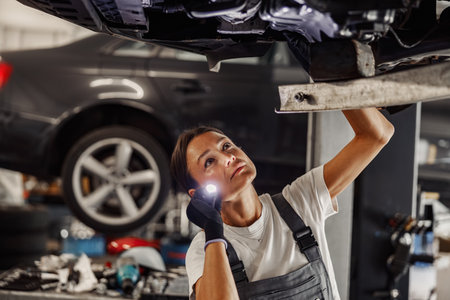A skilled female mechanic inspects a cars underbody with a flashlight in a garageの写真素材