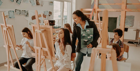Hispanic teacher guiding diverse young students painting on easels inside a bright art studio