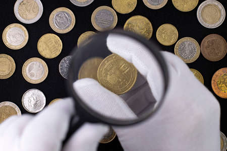 A hand in a white glove holds a coin and a magnifying glass against the background of a pattern of coins on a black cloth.の写真素材