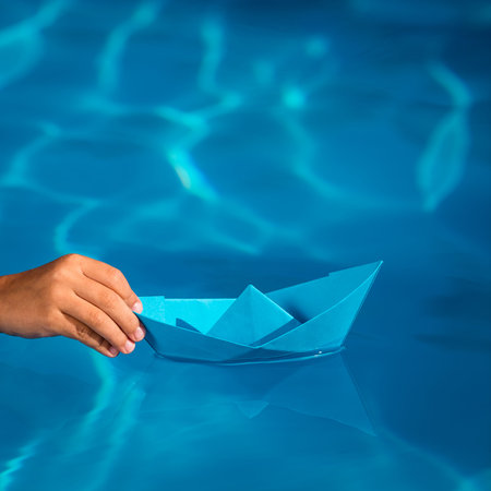 Child holding blue paper sailing boat on water background. Summer vacation and travel concept