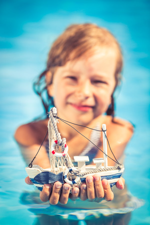 Happy child holding toy sailing boat in hands. Kid playing in swimming pool. Summer vacation and travel conceptの写真素材