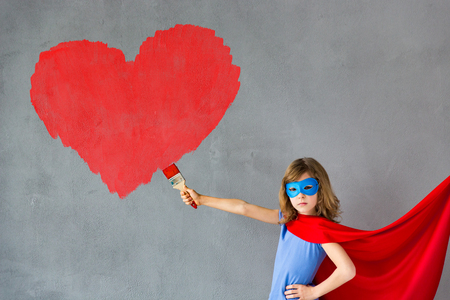 Happy child painting big red heart on the wall. Funny superhero girl playing at home. Valentines day card. Renovation and design conceptの写真素材
