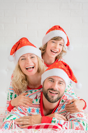 Happy family wearing Santa Claus hats lying on bed. Mother; father and child having fun in Christmas time. Man, woman and kid relaxing at home. Winter holiday Xmas and New Year conceptの写真素材