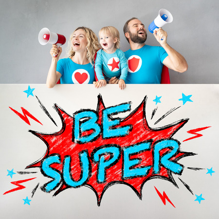 Family of superheroes playing at home. Father, mother, daughter and son holding banner blank. Parents with children pretend to be a super heroesの写真素材