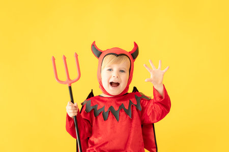Funny child dressed devil costume against yellow background. Happy Halloween holidays concept
