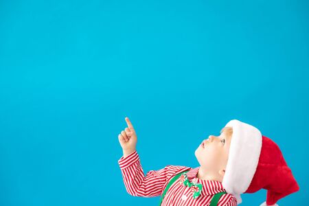 Serious child dressed Santa Claus hat against blue background with copy space. Baby boy point fingers. Christmas holiday conceptの写真素材