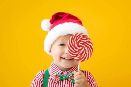 Happy child holding lollipop in hand against yellow background. Portrait of funny kid dressed Santa Claus hat. Christmas holiday conceptの写真素材
