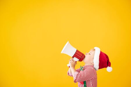 Happy child wearing Santa Claus costume speaking by megaphone. Portrait of funny kid against yellow backgroundの写真素材
