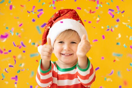 Portrait of happy child showing thumbs up. Funny kid dressed Santa Claus hat against yellow background. Christmas holiday conceptの写真素材